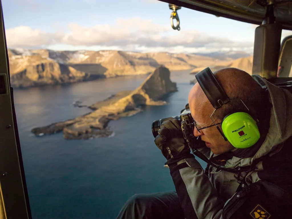 Photo showing Heiner Lamprecht sitting in a flying helicopter next to an open door. He holds a camera to shoot some aerial photos of the coast of Faroer Islands. Photo courtesy of Peter Cox Photo showing Heiner Lamprecht sitting in a flying helicopter next to an open door. He holds a camera to shoot some aerial photos of the coast of Faroer Islands. Photo courtesy of Peter Cox